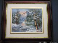 Framed winter landscape oil painting with snow-covered trees, river, and mountains, showing full view including frame and signature.