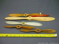 Five vintage model airplane propellers laid flat on a gray surface with measuring tape showing size.