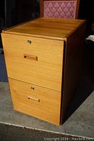 Full view of the filing cabinet showing two drawers, teakwood veneer, and horizontal wooden handles. Cabinet sits on floor near a pink patterned chair.