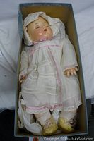 Full view of vintage doll lying in box wearing white dress, bonnet, socks, and yellow shoes.