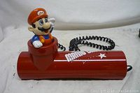 Side angle view showing red pipe-shaped base with Mario figurine and coiled cord