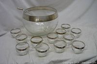 Full view of large punch bowl with ladle and 12 matching glass cups, all with silver rims.