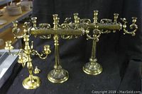 Three brass candelabra shown together on a table with black cloth background. Two candelabra have multiple arms and detailed bases; one features animal figures. The third is smaller with three arms.