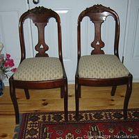 Photo showing pair of Victorian walnut dining chairs with carved floral top rails, upholstered seats, set on hardwood floor with patterned area rugs.