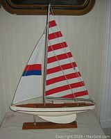 Front view of wooden model sail boat showing red and white striped main sail and jib sail with blue and red stripe, mounted on wooden stand.