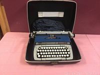Open view of blue Smith Corona Classic 12 manual typewriter inside hard carrying case with dust cover.