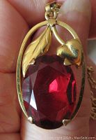Close-up of pendant showing the deep red faceted oval stone set with prongs and decorative heart and leaf gold-tone motif