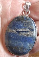 Oval lapis lazuli pendant held in hand showing front polished stone surface with natural inclusions and silver-tone setting and bail.