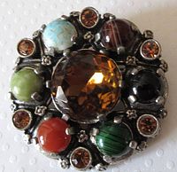 Top view showing the round brooch with a large central faceted amber glass stone, 8 surrounding multi-colored glass stones resembling agates and quartz, and smaller amber rhinestones in between.
