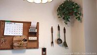 Showing cork board with November 2019 calendar, wicker basket with pens, wooden organizer with drawers and papers, brass decorative kitchen tools on wall, and faux flower arrangement in macrame pot.