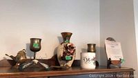 Shelf with the 10 inch floral vase center, pair of brass wine glasses flanking it a small brass plate and mini coal scuttle, hunt mug, and Norman Rockwell collector plate on easel with COA