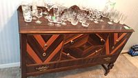 Front angled view of vintage wooden china cabinet with glassware on top, showing decorative wood grain pattern, doors, and drawer fronts.