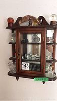 Wooden wall cabinet with glass door displaying a collection of glass decor items such as animals, thimbles, mini teapot, snow globe, and other unique glass pieces.