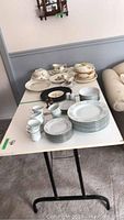 View of white melamine table with dishes and cups placed on top, showing the entire table surface and collapsible black legs; some damage visible on edges and corners.