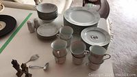 Photo showing stacked large dinner plates, side plates, bowls, cups, and ceramic salt and pepper shakers along with brass salt and pepper shakers and small brass serving utensils on a table.