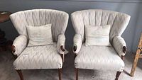Pair of matched upholstered armchairs with cushions and wooden legs and arms shown from front angle on carpeted floor.