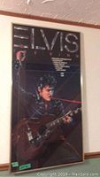 Full view of a framed Elvis Presley poster with the singer playing guitar, dark background
