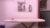 Ironing board with beige floral padded cover, Proctor Silas steam iron mounted on the wall, metal wall rack with clothes shaver and starch bottle above.