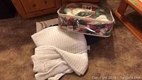 Photo of two white blankets, one folded green blanket, and a multi-colored floral patchwork twin size comforter in a clear zippered case on carpeted floor.