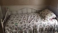 White metal framed day bed made with vertical and curved bars, seen from an angle with floral quilt and pillow on top.