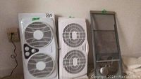 Three window fan units standing upright against a wall, including two dual white plastic case fans and one screen fan with metal frame, showing cords and power plugs.