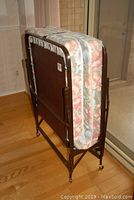 Folded cot bed with floral mattress, metal frame, and casters shown vertically in indoor setting.