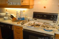 Full kitchen countertop with stacked ceramic plates, bowls, metal utensils, and plastic storage bowls lined up on countertop and stove.