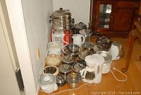 Wide view showing assortment of small kitchen appliances and cookware including coffee maker, kettles, rice cooker, pots and pans, steamer, and pressure cooker all grouped on floor