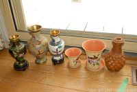 All six items arranged in a line by a window, showing three cloisonne vases, two floral ceramic planters, and one woven vase.