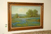 Framed landscape painting showing field with blue flowers, cabin, trees under cloudy sky; wooden frame with some scratches.