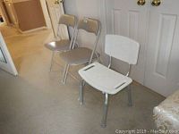 View of two grey folding chairs and one white bath seat placed on carpeted floor near closed doors.