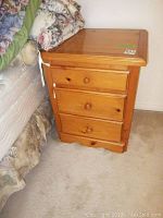 Front view of pine 3 drawer nightstand beside bed, showing drawer knobs and some scratches on wood surface.