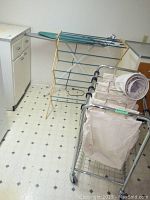 Photo showing the ironing board resting against the wall, foldable drying rack positioned on the floor, and the metal rolling laundry sorter with three canvas bags.