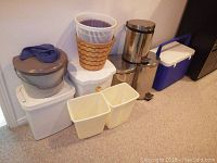 Photo showing ten assorted trash cans and one large blue Coleman cooler against a wall indoors.