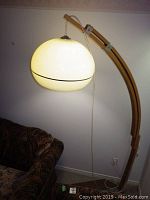 Floor lamp lit, showing curved wood base and white plastic large rounded shade against wall near a patterned couch.