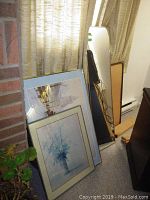 Multiple framed art prints and a mirror leaning against a wall near a curtain and brick wall. Prints vary in size; largest approx 24x36. Frames mostly wood or metal in neutral tones.