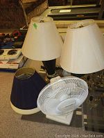 Photo shows two ceramic base table lamps with white shades, one dark blue lampshade, and a white Duracraft brand table fan.