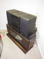 Full view of Kenwood stereo system with speakers placed on top of wood grain stereo cabinet base. Includes remote control on cabinet and visible wiring.