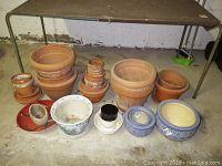 Overall view of assorted plant pots including terra cotta and ceramic hand painted pots under a table; some pots are stacked inside others, showing sizes and condition.