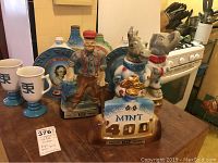 Photo of eight Jim Beam collectible ceramic flasks and two white stemmed mugs on a wooden table near a kitchen stove.