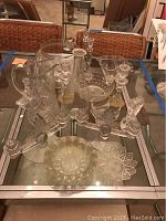 Glass candlesticks with winged designs alongside clear glass bowls and pitcher on glass table.