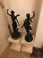 Pair of bronze dancing lady statuettes on separate pedestals, one with green marble base and the other with glass top, displayed against a cream wall corner.