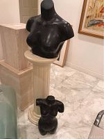 Large matte black female torso sculpture displayed on cream-colored fluted pedestal indoors on marble floor.