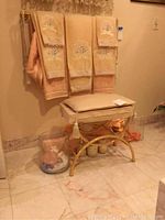 Peach towels hanging on gold towel rod above padded stool with gold frame, stone bathroom set on floor, glass trash can, and acrylic holder visible
