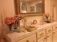 Overview of items on marble bathroom countertop, gold towel holders, faux flower vases, candle holders, perfume bottles, clock radio