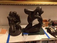 Two carved wood statues side by side on a shelf, includes smaller statue of two seated embracing figures and larger abstract statue on rectangular base.