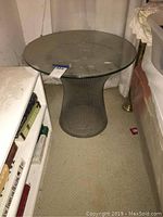 Side table with metal wire pedestal base and glass top showing soiling, placed next to bookshelf and couch.