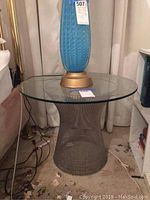 Glass top pedestal side table with wire metal base, showing soiling on the base and carpet below. Blue lamp on top for size context, lamp not included.