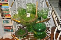 Green glass plates, pitchers, vases and bowls arranged on a triangular metal rack with gold accents and a wood floor background. Books seen to the left.