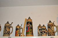 Six German-style beer steins arranged on shelf, captured with ruler for scale, highlighting pewter lids, ornate handles, and relief artwork.